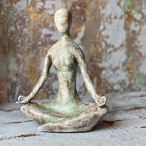 Puede incluir: Una escultura de una figura de yoga sentada en posici&oacute;n meditativa. La figura es de color verde y gris apagado, con los brazos extendidos y las manos apoyadas en las rodillas. La escultura est&aacute; sobre una superficie texturizada.