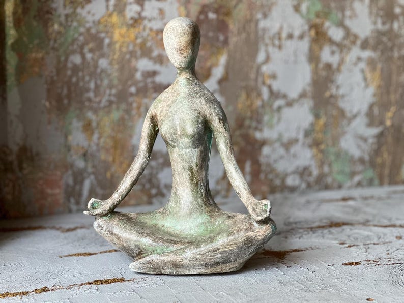 Puede incluir: Una escultura de una figura de yoga sentada en posici&oacute;n meditativa. La figura es de color verde y gris apagado, con los brazos extendidos y las manos apoyadas en las rodillas. La escultura est&aacute; sobre una superficie texturizada.