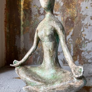 Puede incluir: Escultura de bronce envejecido de una persona en postura de yoga sentada. La figura tiene un rostro liso y est&aacute; sentada con las piernas cruzadas y los brazos extendidos. La escultura tiene detalles en verde y marr&oacute;n.