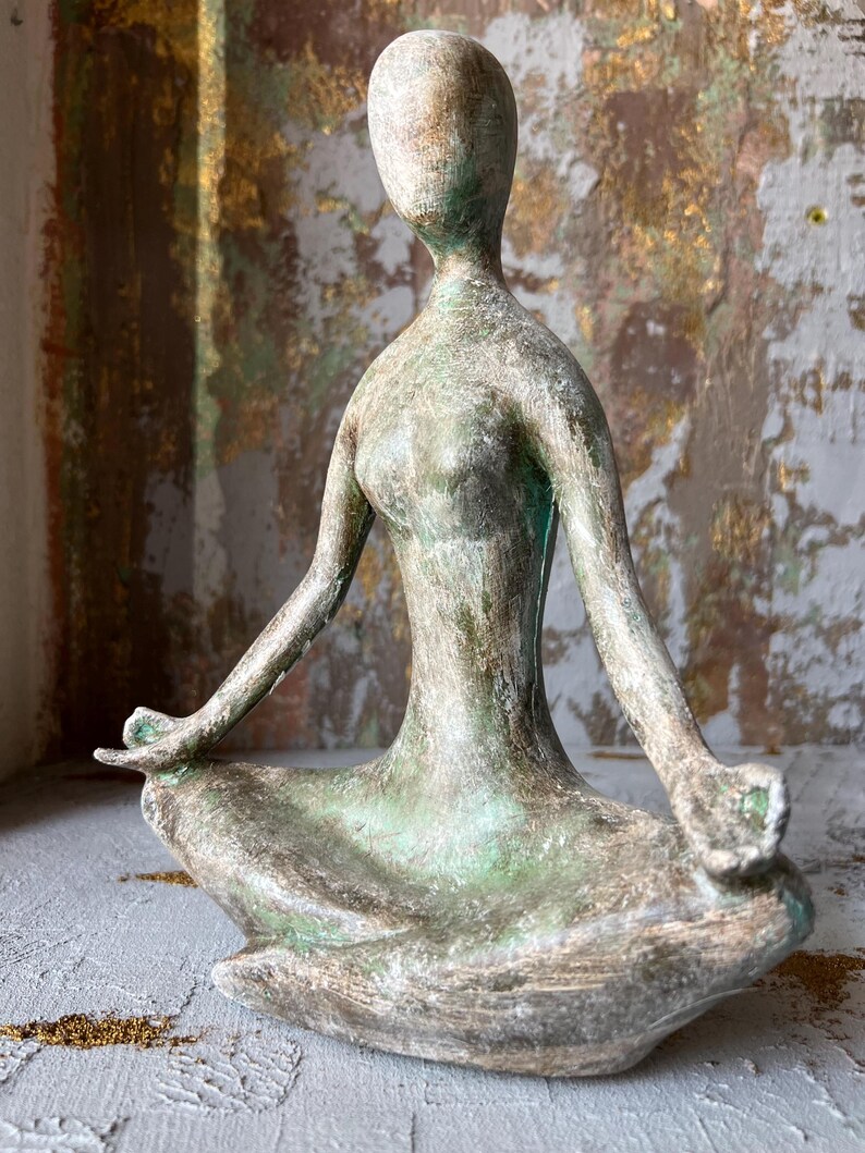 Puede incluir: Escultura de bronce envejecido de una persona en postura de yoga sentada. La figura tiene un rostro liso y est&aacute; sentada con las piernas cruzadas y los brazos extendidos. La escultura tiene detalles en verde y marr&oacute;n.