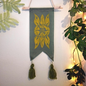 Peut inclure: Une bannière vert sauge avec un motif floral jaune et deux glands verts. La bannière est suspendue à une cheville en bois et à une corde crème. La bannière est accrochée à un mur blanc.