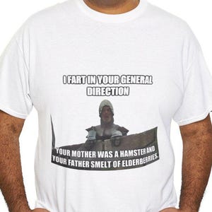 May include: White t-shirt with a humorous graphic. The graphic features text that reads "I FART IN YOUR GENERAL DIRECTION" above an image of a medieval knight. Below the image, the text continues with "YOUR MOTHER WAS A HAMSTER AND YOUR FATHER SMELT OF ELDERBERRIES."