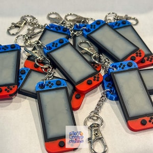 Könnte beinhalten: Eine Gruppe von roten und blauen Nintendo Switch Schlüsselanhängern mit einer silbernen Kette und einem silbernen Verschluss. Die Anhänger sind in Form der Nintendo Switch Konsole geformt und haben einen leeren Bildschirm.