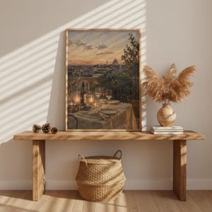 Pode incluir: Uma impress&atilde;o de arte de parede vertical emoldurada retratando uma cena de terra&ccedil;o ao p&ocirc;r do sol com uma mesa posta para jantar, apresentando ta&ccedil;as de vinho, uma garrafa e pratos. A obra est&aacute; exposta numa mesa de consola de madeira com um vaso de ervas.