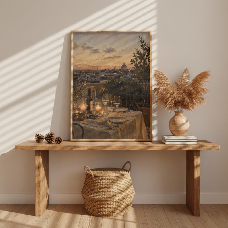 Pode incluir: Uma impress&atilde;o de arte de parede vertical emoldurada retratando uma cena de terra&ccedil;o ao p&ocirc;r do sol com uma mesa posta para jantar, apresentando ta&ccedil;as de vinho, uma garrafa e pratos. A obra est&aacute; exposta numa mesa de consola de madeira com um vaso de ervas.