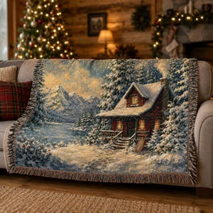 Könnte beinhalten: Eine gewebte Wohndecke mit einer Winterlandschaft, die eine rote Hütte inmitten verschneiter Tannen und Berge zeigt. Das Motiv umfasst einen zugefrorenen See, sanftes goldenes Licht aus den Hüttenfenstern und einen Rand mit Fransen.