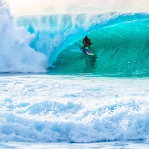 May include: A surfer rides a wave, the water is a vibrant turquoise blue, and the wave is breaking into white foam.