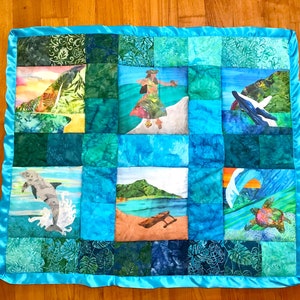 May include: A rectangular quilt featuring a bright turquoise satin border and a patchwork design of various blue and green fabrics. The quilt displays printed scenes of a hula dancer, a breaching whale, a dolphin, a sea turtle, and a beach landscape.