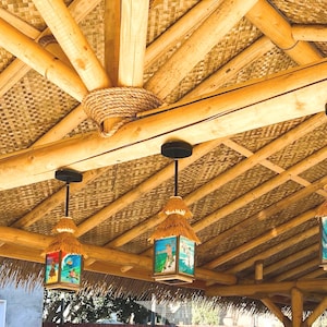 May include: Four square pendant lights with thatched roofs and colorful surf-themed panels hang from a bamboo ceiling structure. Each light features a black metal mount and a rectangular frame displaying illustrations of ocean waves and surfers.