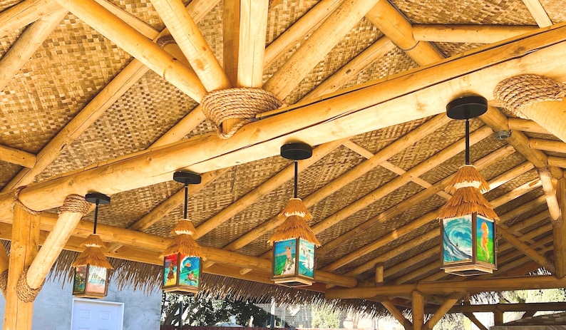 May include: Four square pendant lights with thatched roofs and colorful surf-themed panels hang from a bamboo ceiling structure. Each light features a black metal mount and a rectangular frame displaying illustrations of ocean waves and surfers.