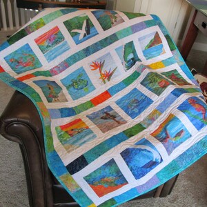 May include: A patchwork quilt featuring a grid of square fabric panels with various tropical scenes, including sea turtles, whales, flowers, and coastal landscapes, framed by white borders and vibrant blue, green, and orange patterned fabric strips.