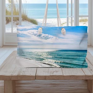 May include: A rectangular throw pillow featuring a photographic print of a sunny beach scene with turquoise ocean waves, white sand, and a bright blue sky with wispy clouds and two small white birds flying in the upper center of the image.