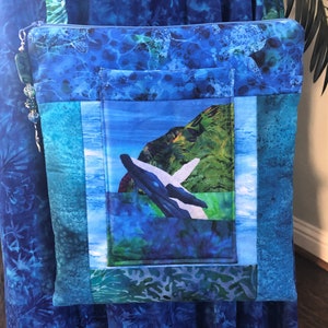 May include: A square fabric crossbody bag featuring a blue batik-style print with sea turtles. The front includes a central pocket displaying a whale breaching near a green island. A blue zipper closure is accented with a beaded silver charm.
