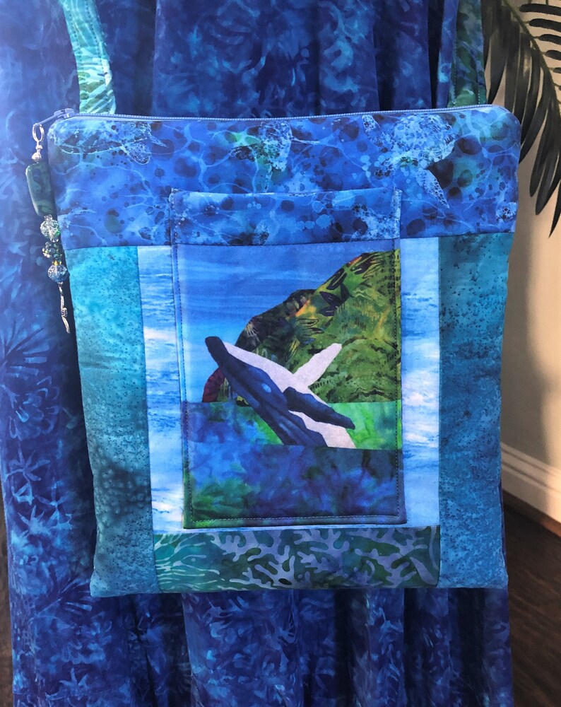 May include: A square fabric crossbody bag featuring a blue batik-style print with sea turtles. The front includes a central pocket displaying a whale breaching near a green island. A blue zipper closure is accented with a beaded silver charm.