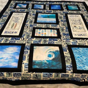 May include: A patchwork quilt featuring a beach and surfing theme with blue, white, and tan colors. It includes rectangular panels of ocean waves, surfboards, and text such as Low Tides Good Vibes, Sand Sun Surf Fun, and Cruisin Surfin Chillin.