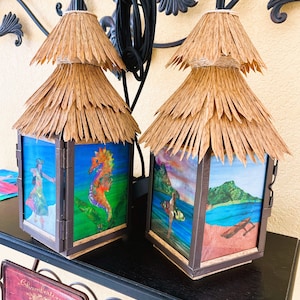 May include: Two square lanterns with dark brown frames and tiered, natural-colored straw roofs. Each lantern features four glass panels decorated with colorful illustrations, including a seahorse, a hula dancer, a surfer, and a coastal landscape.