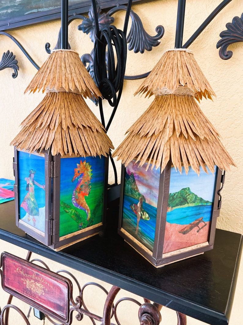May include: Two square lanterns with dark brown frames and tiered, natural-colored straw roofs. Each lantern features four glass panels decorated with colorful illustrations, including a seahorse, a hula dancer, a surfer, and a coastal landscape.