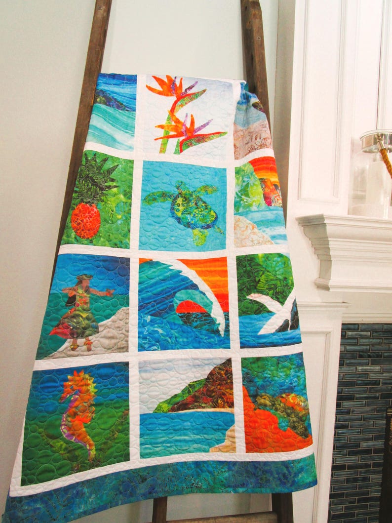 May include: A quilt featuring a grid of twelve square panels with vibrant tropical imagery, including a sea turtle, a seahorse, a hula dancer, a pineapple, a wave, and bird of paradise flowers, all separated by white fabric borders.