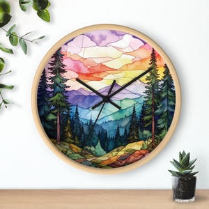 May include: A round wall clock with a light wood frame featuring a stained glass style illustration of a mountain landscape with pine trees, a colorful sunset sky in shades of pink, orange, and yellow, and black clock hands.