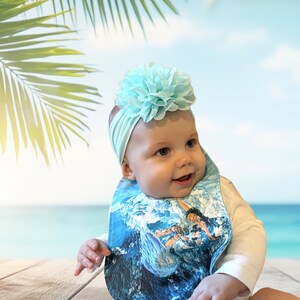 May include: A baby wearing a white onesie with a blue and white patterned bib. The bib features a surfer riding a wave. The baby is wearing a light blue headband with a large light blue flower on top.
