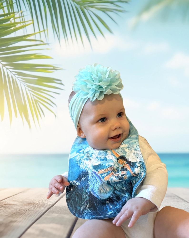 May include: A baby wearing a white onesie with a blue and white patterned bib. The bib features a surfer riding a wave. The baby is wearing a light blue headband with a large light blue flower on top.