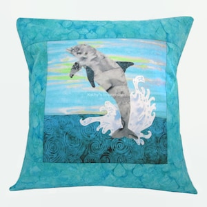 May include: A square throw pillow featuring a central panel with a gray dolphin leaping from dark blue swirling water against a light blue sky. The text Kathy's Impressions is printed in the center. The border is a textured turquoise fabric.
