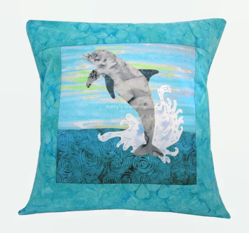 May include: A square throw pillow featuring a central panel with a gray dolphin leaping from dark blue swirling water against a light blue sky. The text Kathy's Impressions is printed in the center. The border is a textured turquoise fabric.