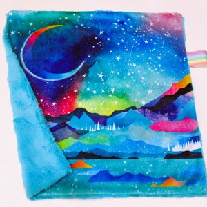 以下が含まれることがあります： 天体デザインの正方形のベビーブランケット。三日月、星空、青、緑、ピンク、オレンジの色合いの山々の鮮やかな水彩画のシーンが特徴です。明るい青色のふわふわの縁がブランケットを囲み、虹色のリボンが付いています。