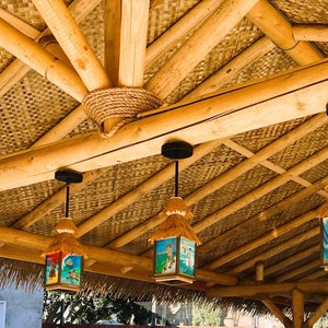 May include: Four hanging pendant lights with thatched roofs and square glass panels featuring colorful surf-themed illustrations, suspended from a natural bamboo ceiling structure with woven matting and thick rope accents at the joints.