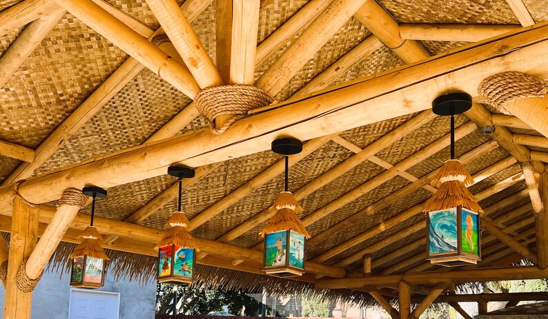 May include: Four hanging pendant lights with thatched roofs and square glass panels featuring colorful surf-themed illustrations, suspended from a natural bamboo ceiling structure with woven matting and thick rope accents at the joints.