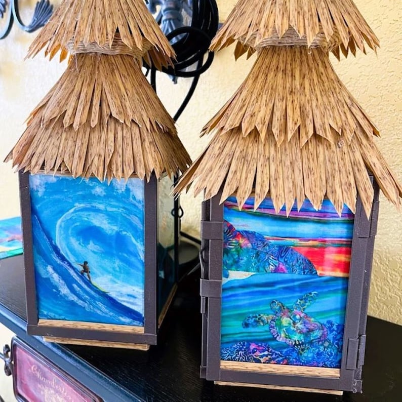 May include: Two brown and black tiki-style lanterns with thatched roofs. The lantern on the left has a blue and white image of a surfer riding a wave. The lantern on the right has a blue, green, and purple image of a sea turtle swimming in the ocean.
