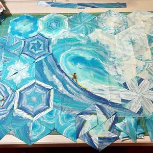 May include: A collection of fabric hexagons arranged to form a large ocean wave scene. The quilt blocks feature shades of blue, teal, and white, depicting a crashing wave with a small surfer on a yellow board in the center of the composition.