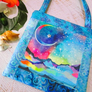 以下が含まれることがあります： 天体デザインの正方形トートバッグ。三日月と星が描かれた夜空の水彩画が特徴です。バッグは青い模様の生地でトリミングされ、お揃いのハンドルが付いています。