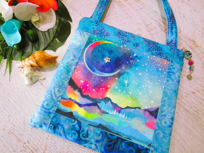 以下が含まれることがあります： 天体デザインの正方形トートバッグ。三日月と星が描かれた夜空の水彩画が特徴です。バッグは青い模様の生地でトリミングされ、お揃いのハンドルが付いています。