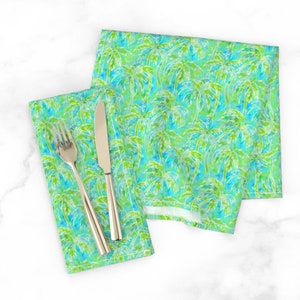 May include: Two green and blue napkins with a white palm tree pattern. The napkins are folded and placed on a white surface with a silver fork and knife.