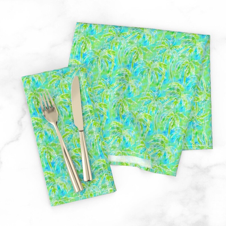 May include: Two green and blue napkins with a white palm tree pattern. The napkins are folded and placed on a white surface with a silver fork and knife.