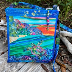 May include: A blue and green fabric crossbody bag with a colorful sea turtle design and a beaded strap. The bag has a patchwork design with a sunset scene and floral patterns.