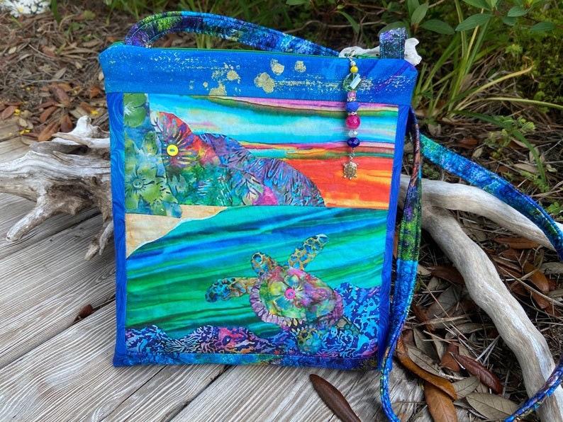 May include: A blue and green fabric crossbody bag with a colorful sea turtle design and a beaded strap. The bag has a patchwork design with a sunset scene and floral patterns.