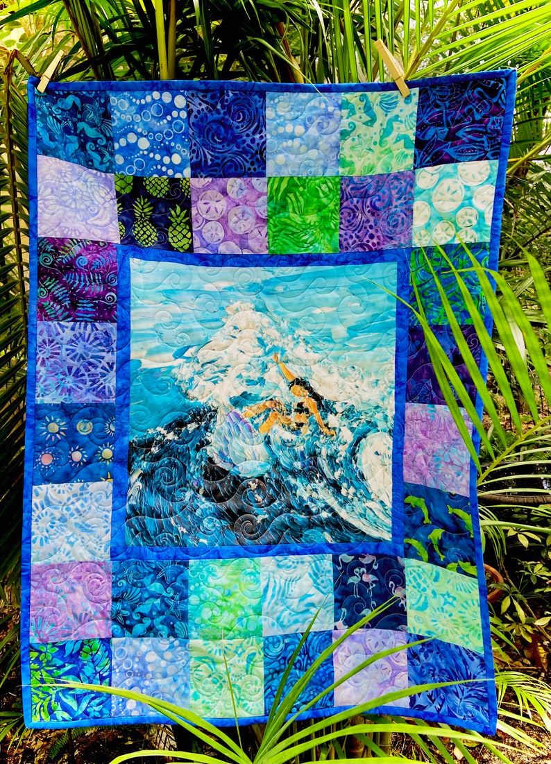May include: A blue and purple patchwork quilt with a central panel depicting a surfer riding a wave. The quilt is made of various fabrics with different patterns, including pineapples, seashells, and fish.