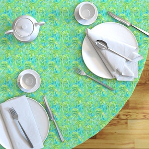 May include: A round tablecloth with a tropical palm tree pattern in shades of green, blue, and white. The tablecloth is set with a white teapot, a white saucer with a cup, a white plate with a white napkin and silverware, and a white saucer with a cup.