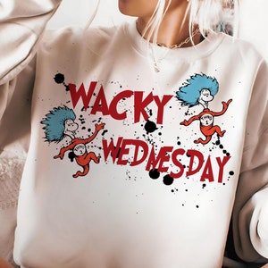 May include: Cream-colored sweatshirt with the words "WACKY WEDNESDAY" in red, featuring cartoon characters with blue hair and red bodies. Black ink splatters accent the design. A casual, comfortable style.