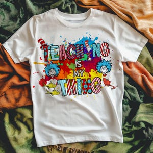 May include: White t-shirt with the text "TEACHING IS MY THING" in colorful, patterned letters. The design includes cartoon characters and a vibrant paint splatter background, creating a playful and educational theme.