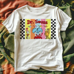 Puede incluir: Camiseta blanca con un dise&ntilde;o colorido de personajes de Dr. Seuss, incluyendo el Gato en el Sombrero, Thing 1 y Thing 2, y el Lorax. El dise&ntilde;o incluye el texto "Dr. Seuss" en letras rojas. El dise&ntilde;o est&aacute; enmarcado por patrones de cuadros blancos y negros.