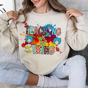 May include: Beige sweatshirt with the text "TEACHING IS MY THING" in colorful, patterned letters. The design includes Dr. Seuss-inspired characters and paint splatters in various colors. The sweatshirt is a casual, long-sleeved garment.