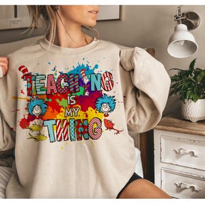 May include: Beige sweatshirt with the phrase "TEACHING IS MY THING" in colorful, whimsical lettering. The design includes illustrations of characters and splashes of paint, creating a playful and educational theme.