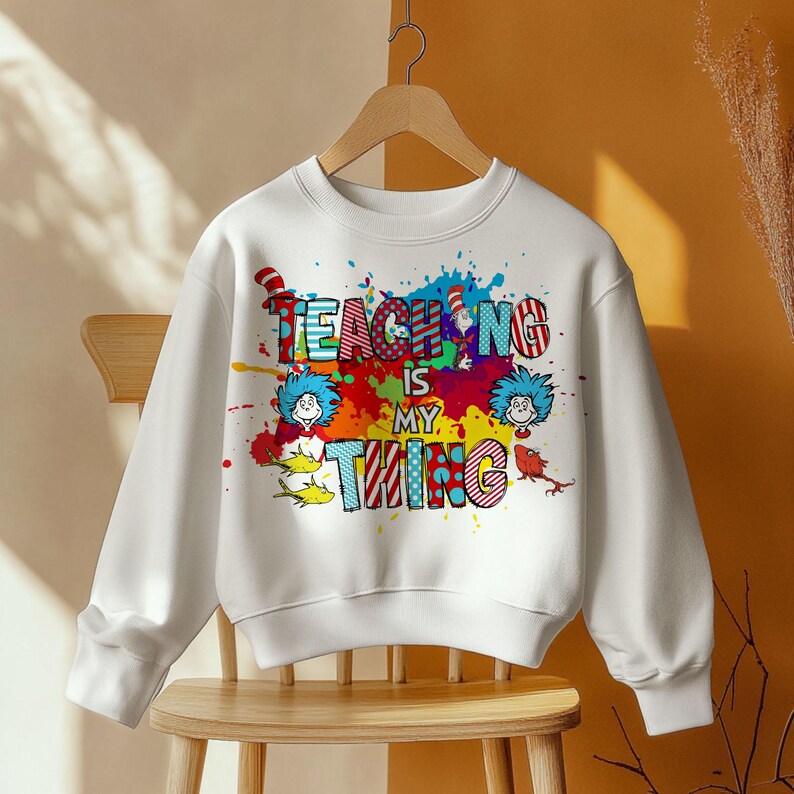 May include: White sweatshirt with the phrase "Teaching is my thing" in colorful, whimsical lettering. The design includes cartoon characters and paint splatters, creating a playful and educational theme. The sweatshirt is displayed on a wooden hanger.