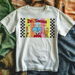 Puede incluir: Camiseta blanca con un gr&aacute;fico colorido de personajes del Dr. Seuss. El dise&ntilde;o incluye el texto "Dr. Seuss" e ilustraciones del Gato en el Sombrero, Thing 1 y Thing 2, y el Lorax. El gr&aacute;fico est&aacute; enmarcado con bordes a cuadros en blanco y negro.