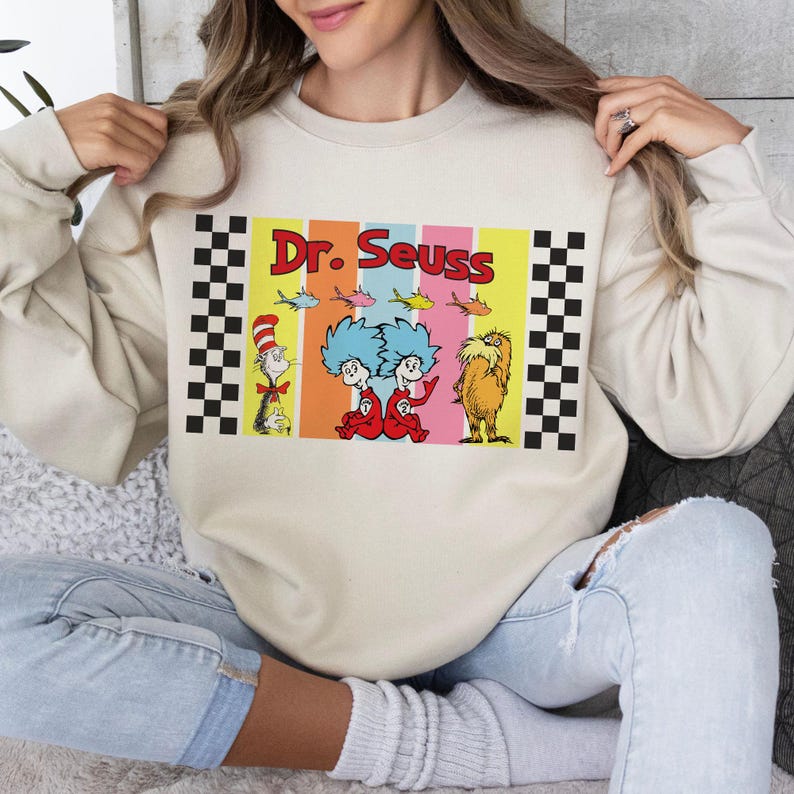 Puede incluir: Sudadera beige con un gr&aacute;fico colorido de personajes de Dr. Seuss, incluyendo el Gato en el Sombrero, Thing 1 y Thing 2, y el Lorax. El gr&aacute;fico est&aacute; enmarcado con patrones de cuadros blancos y negros. Las palabras "Dr. Seuss" est&aacute;n impresas en rojo.