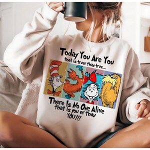 May include: Cream-colored sweatshirt featuring a colorful graphic with characters and text from a children's book. The text reads "Today You Are You" and "There Is No One Alive that is you er than YOU!!!". The graphic includes four cartoon characters.