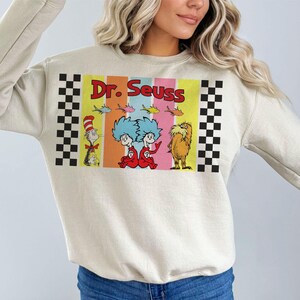 Puede incluir: Sudadera color crema con un colorido gr&aacute;fico de Dr. Seuss. El gr&aacute;fico incluye el texto "Dr. Seuss" en rojo, junto con ilustraciones de personajes como el Gato en el Sombrero, los Cosas y el Lorax. Patrones de cuadros blancos y negros en los lados.